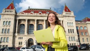 Mosolygó fiatal nő sárga laptoppal egy impozáns, klasszicista stílusú épület előtt.