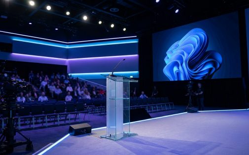 Modern konferenciaterem kék neon megvilágítással, üres szónokpulttal és nagy képernyőkkel a közönség előtt.